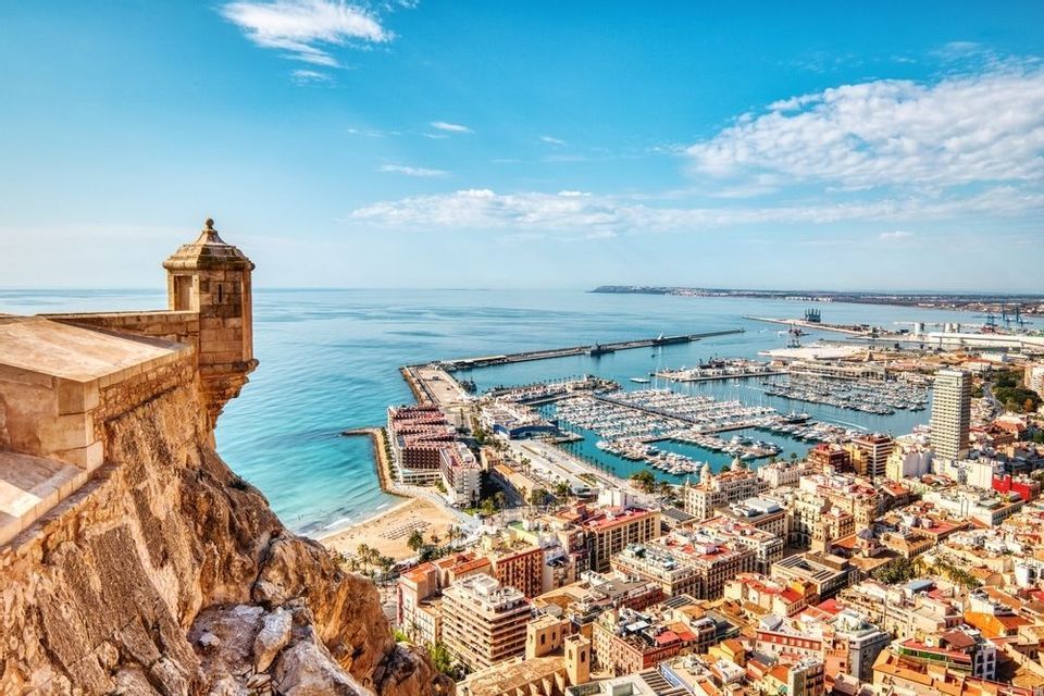 Veduta dall'alto da una fortificazione in pietra che si affaccia su una città costiera con un'ampia marina piena di barche sotto un cielo soleggiato.