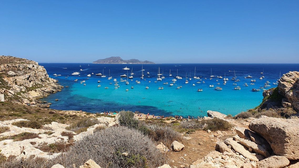Vista aérea de una costa rocosa con una bahía turquesa repleta de numerosos barcos anclados y una isla lejana.