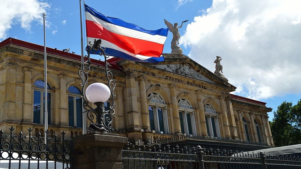 La bandiera del Costa Rica sventola su un'asta di fronte all'ornata facciata in pietra dell'edificio del Teatro Nacional, sotto un cielo azzurro e nuvoloso.