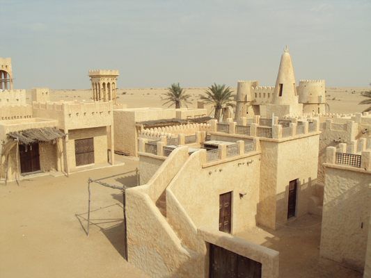 Eine Ansammlung traditioneller, sandfarbener Gebäude und Türme mit Palmen in einer Wüste unter einem diesigen Himmel.