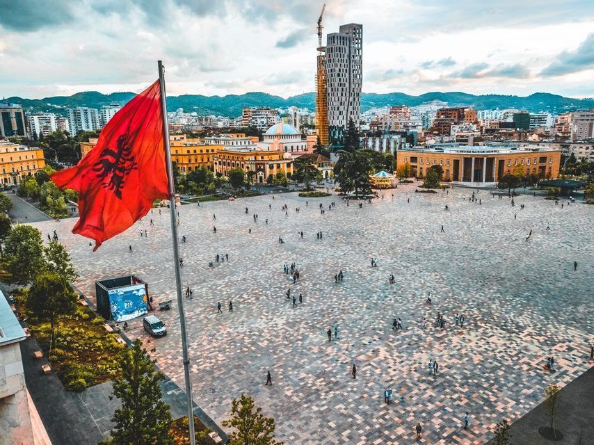 An Albanian flag waves over a large, bustling city square filled with people, with a city skyline in the background.