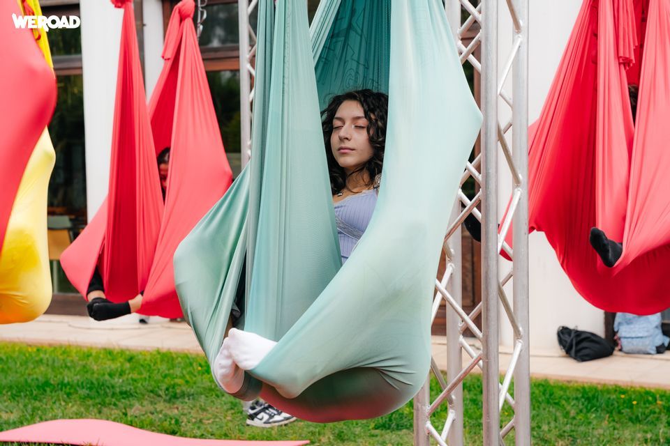 Una giovane donna con gli occhi chiusi si rilassa all'interno di un'amaca yoga aerea verde durante un'attività di gruppo WeRoad all'aperto.