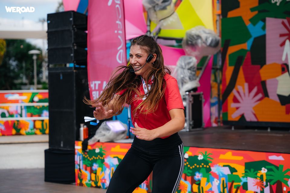 Una donna sorridente con una maglietta rossa e delle cuffie conduce una routine di ballo su un palco all'aperto colorato.