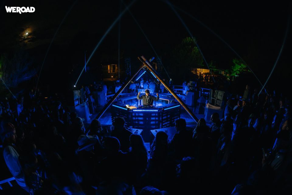 Un DJ si esibisce su un palco triangolare con luci blu per un viaggio di gruppo WeRoad a una festa notturna all'aperto.