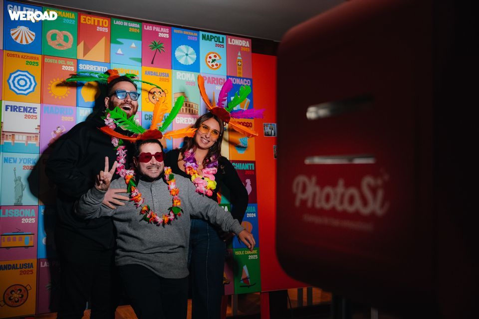 Un gruppo WeRoad di tre persone con accessori festivi in posa per una foto in un fotobooth, con uno sfondo colorato a tema viaggio.