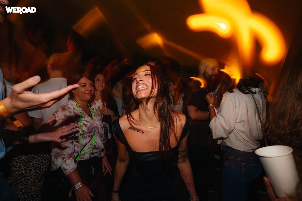 Una donna sorridente balla al centro di una festa di gruppo WeRoad affollata, con luci sfocate dal movimento.