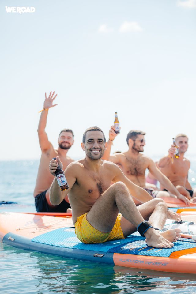 Un viaggio di gruppo WeRoad di uomini seduti su paddleboard in mare, che sorridono e tengono in mano bottiglie di birra.
