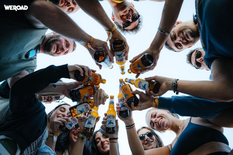 Una vista dal basso di un gruppo WeRoad che brinda con bottiglie di birra sotto un cielo sereno.