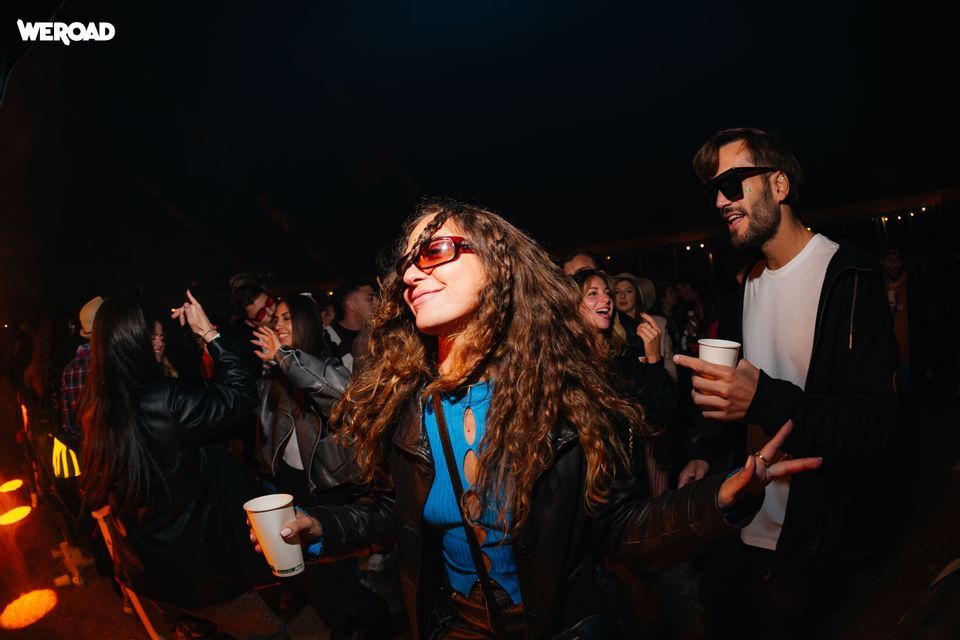 Un gruppo WeRoad in viaggio con persone sorridenti e danzanti a una festa notturna all'aperto, e una donna con gli occhiali da sole in primo piano.