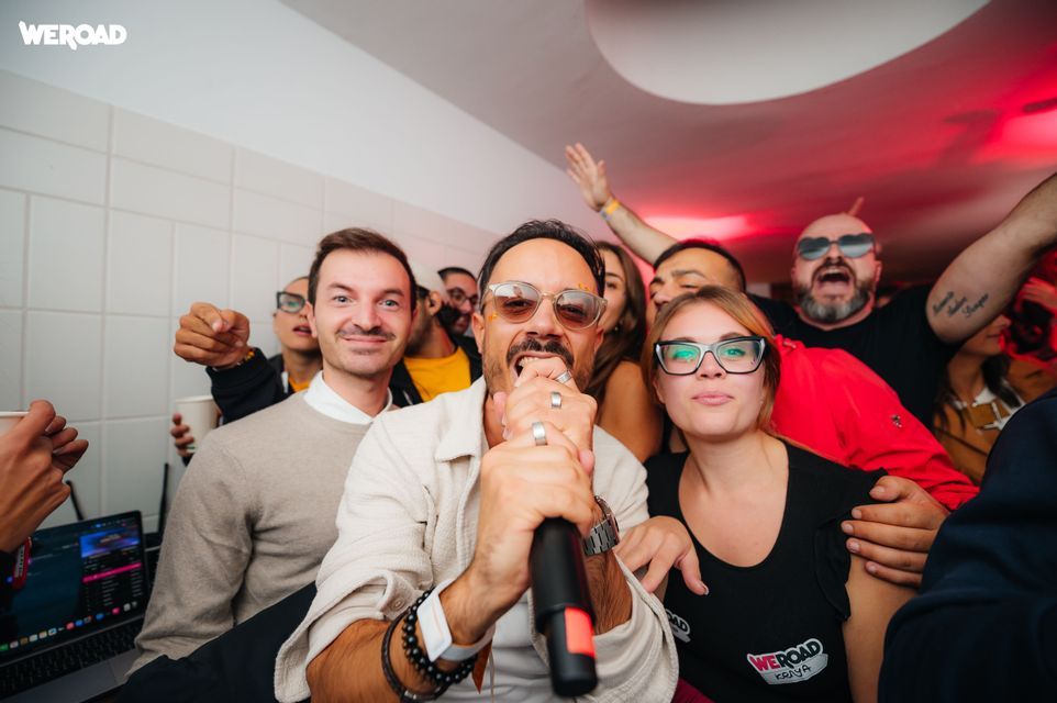 Un uomo canta al microfono mentre un gruppo WeRoad lo acclama e balla attorno a lui a una festa al coperto.