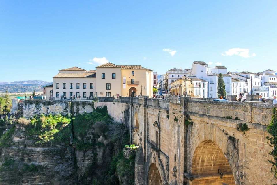 Un grande ponte in pietra con archi che porta a una città di edifici bianchi e beige costruita su una scogliera rocciosa e verde.