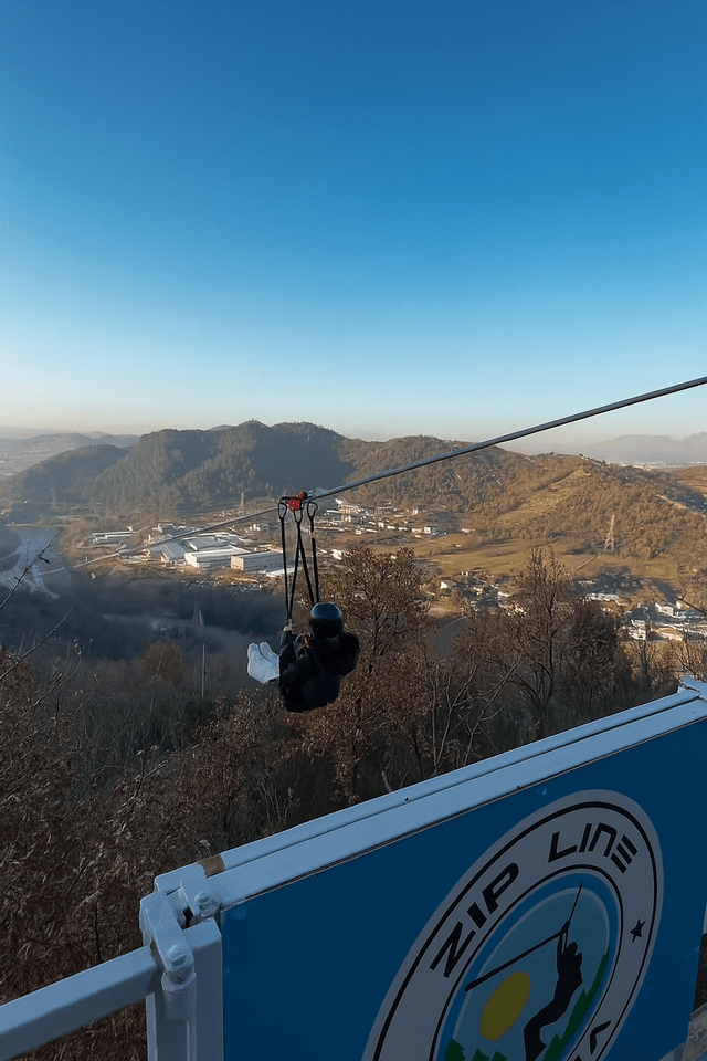 Una persona con un casco scivola lungo una zip line, sospesa su un'ampia valle con colline boscose sotto un cielo azzurro e limpido.