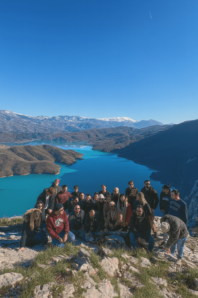 Un viaggio di gruppo WeRoad posa su un punto panoramico roccioso che si affaccia su un lago turchese e montagne innevate in lontananza sotto un cielo azzurro e limpido.