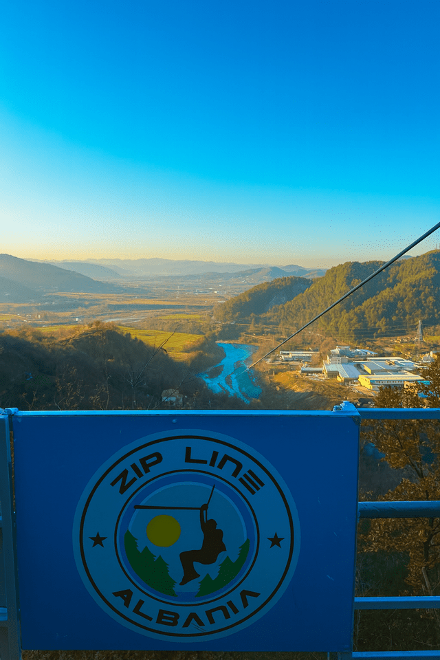 Una vista mozzafiato da una piattaforma di zipline che domina una vasta valle con un fiume turchese e dolci colline sotto un cielo limpido, con il logo 'Zip Line Albania' in primo piano.