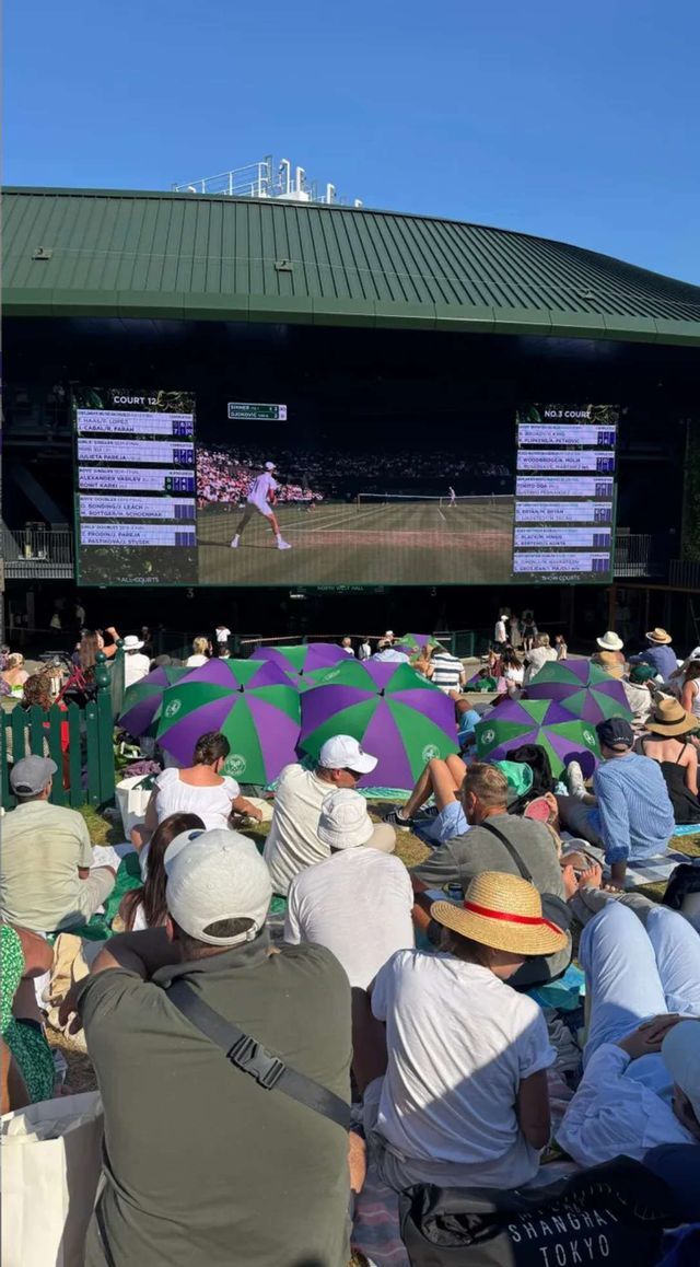 A crowd of people sits on a grassy hill, some with umbrellas, watching a tennis match broadcast on a large outdoor screen.