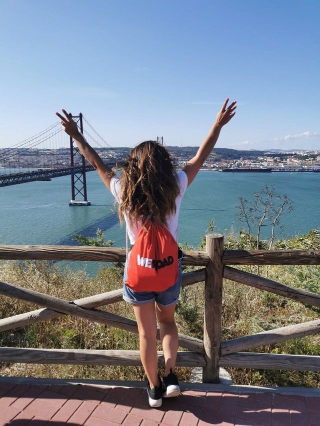Eine von hinten gesehene Frau mit rotem WeRoad-Rucksack hebt die Arme, während sie eine große Brücke und eine Stadt am Wasser überblickt.