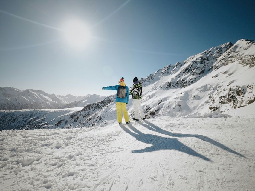 Due persone di un viaggio di gruppo WeRoad su una cima montuosa innevata, che indicano il panorama sotto un sole splendente.