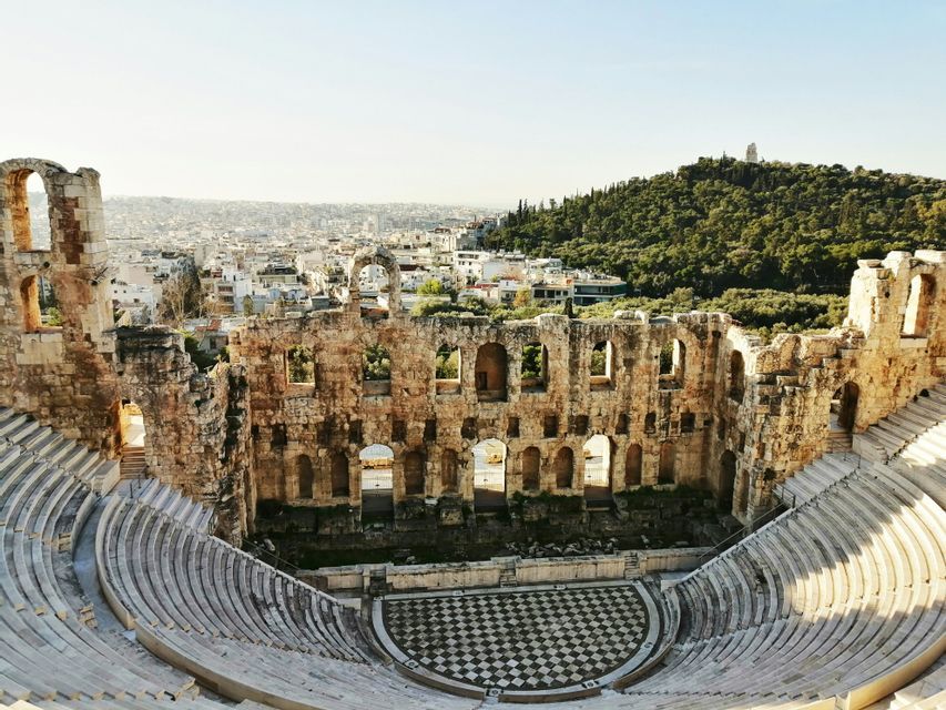 Una vista aérea de un gran y antiguo anfiteatro de piedra en ruinas, con vistas a una ciudad moderna y una colina verde.