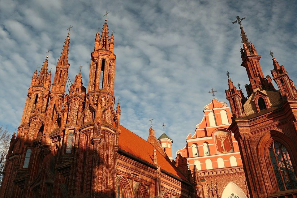 Una vista dal basso di un'ornata chiesa gotica in mattoni rossi, con molteplici guglie, stagliata contro un cielo blu con nuvole bianche.