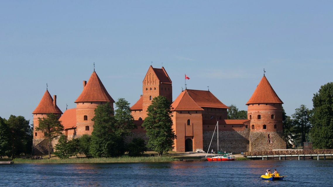 Un grande castello in mattoni rossi con torri coniche si erge sulla riva di un lago, con un pedalò sull'acqua in primo piano.