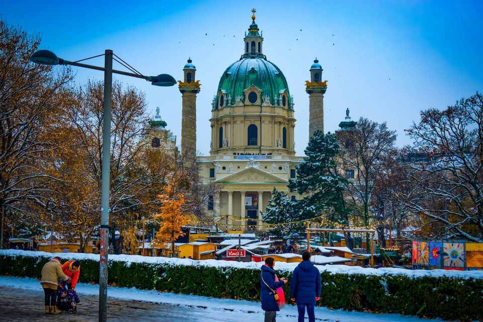 Eine große Kirche mit einer grünen Kuppel dominiert einen verschneiten Park, in dem Leute über einen Wintermarkt schlendern.