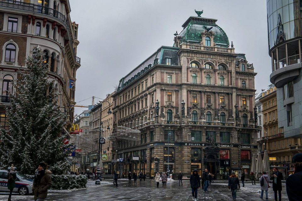 Fußgänger schlendern über einen verschneiten Stadtplatz, geschmückt mit einem großen Weihnachtsbaum und prächtigen historischen Gebäuden, unter einem bedeckten Himmel.
