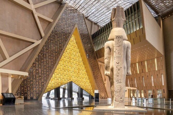 Una colossale statua egizia si erge in una moderna sala museale con pareti triangolari dorate con geroglifici e un soffitto di vetro.