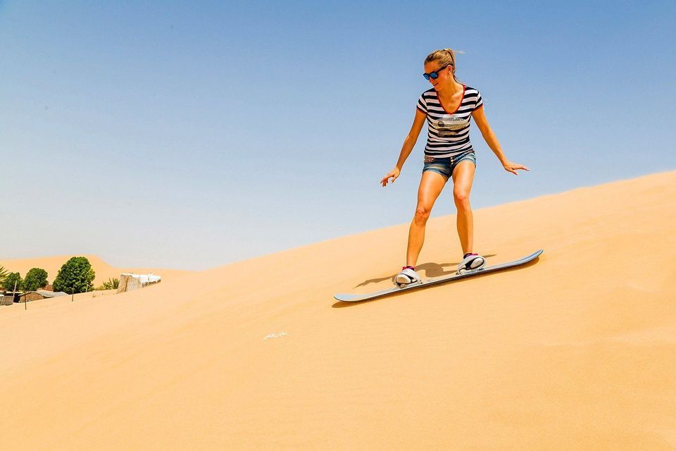 Una donna con una maglietta a righe e occhiali da sole fa sandboarding su una grande duna di sabbia sotto un cielo azzurro e limpido.