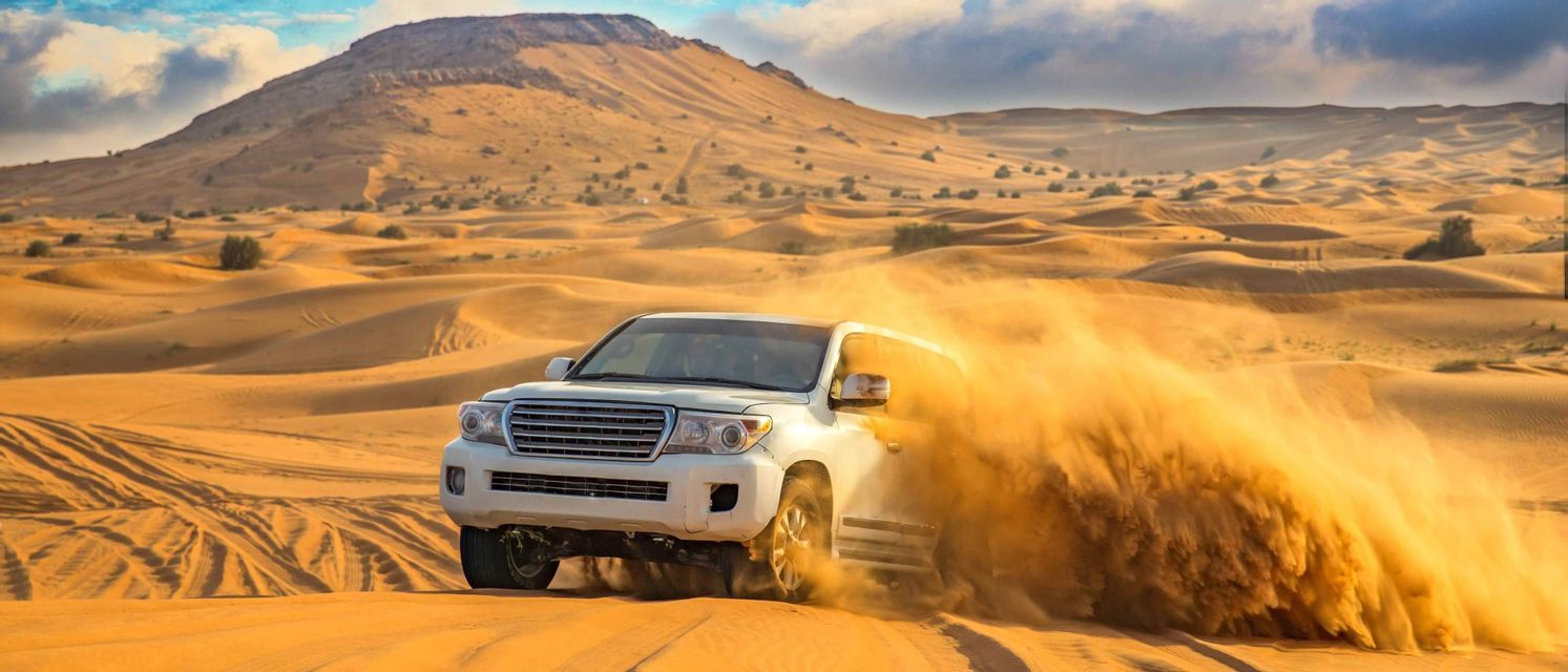 Un SUV bianco solleva una nuvola di sabbia mentre attraversa un vasto deserto con grandi dune sullo sfondo.