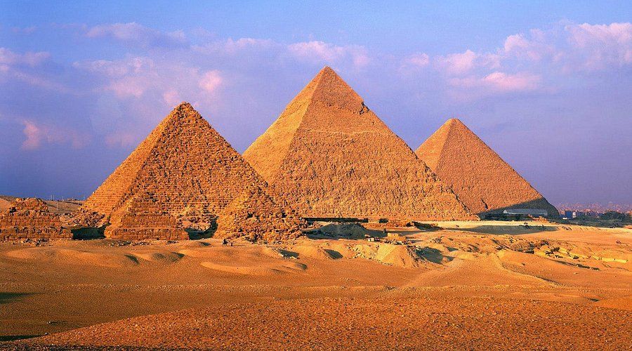 Tre grandi piramidi antiche si ergono in un deserto sabbioso sotto un cielo blu con nuvole sottili al tramonto.