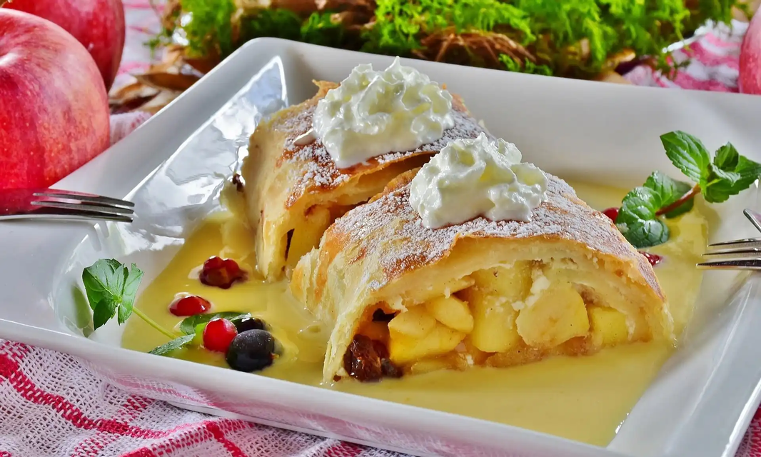 Dos rebanadas de strudel de manzana en un plato blanco, coronadas con crema batida y azúcar glas, servidas sobre un lecho de salsa de vainilla.