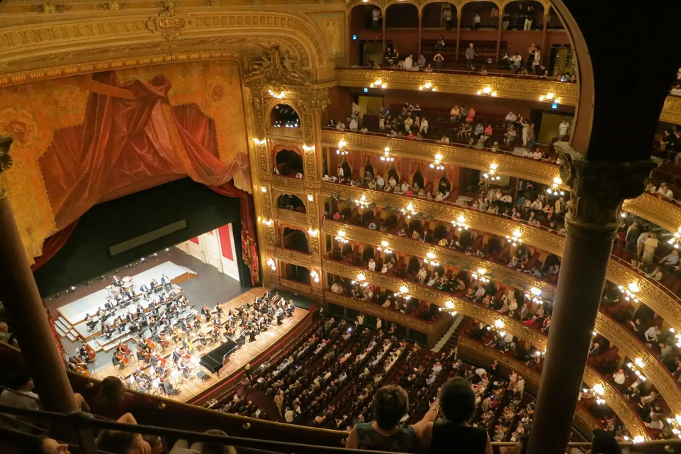 Una vista en picada de una orquesta actuando para el público en un teatro de ópera ornamentado y de varios niveles.