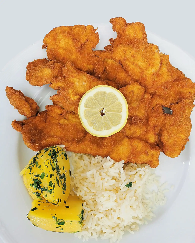 Una milanesa dorada y empanizada en un plato blanco con una rodaja de limón, arroz blanco y patatas con hierbas.