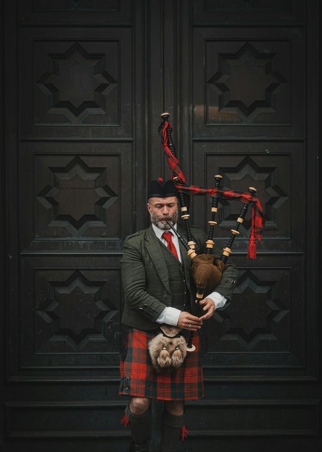 A man in traditional Scottish attire, including a kilt and tweed jacket, holds a set of bagpipes while standing before a large, ornate wooden door.