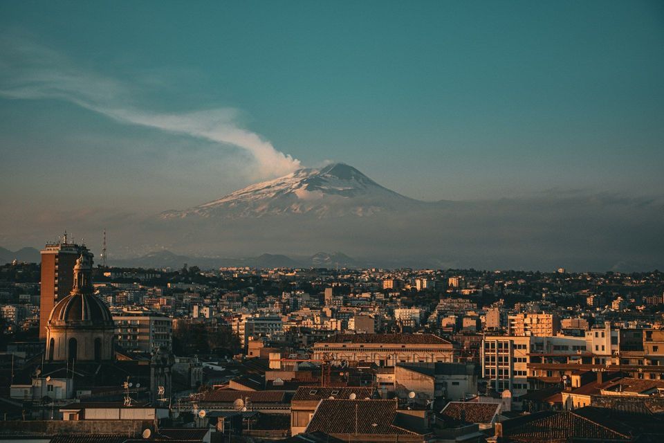 Un paesaggio urbano sotto un vulcano innevato, con un pennacchio di fumo contro un cielo color ottanio.