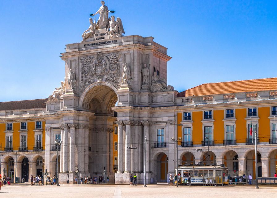 Un tranvía blanco clásico en una plaza soleada frente a un arco de triunfo ornamentado de piedra blanca y un gran edificio amarillo.