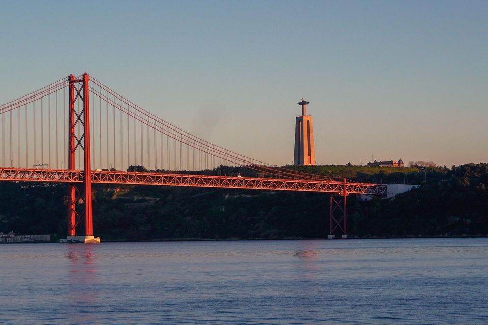 Un puente colgante rojo cruza un cuerpo de agua al atardecer, con un monumento alto visible en una colina verde al fondo.