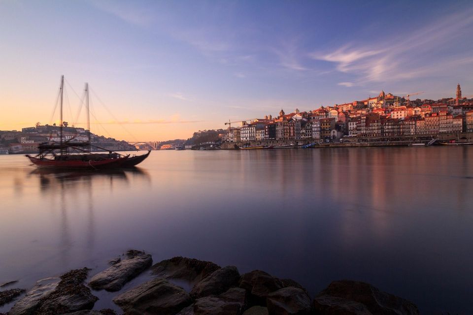 Una barca de madera tradicional en un río tranquilo al atardecer, con los coloridos edificios de una ciudad en la ladera en la orilla opuesta, bajo un cielo rosado y azul.