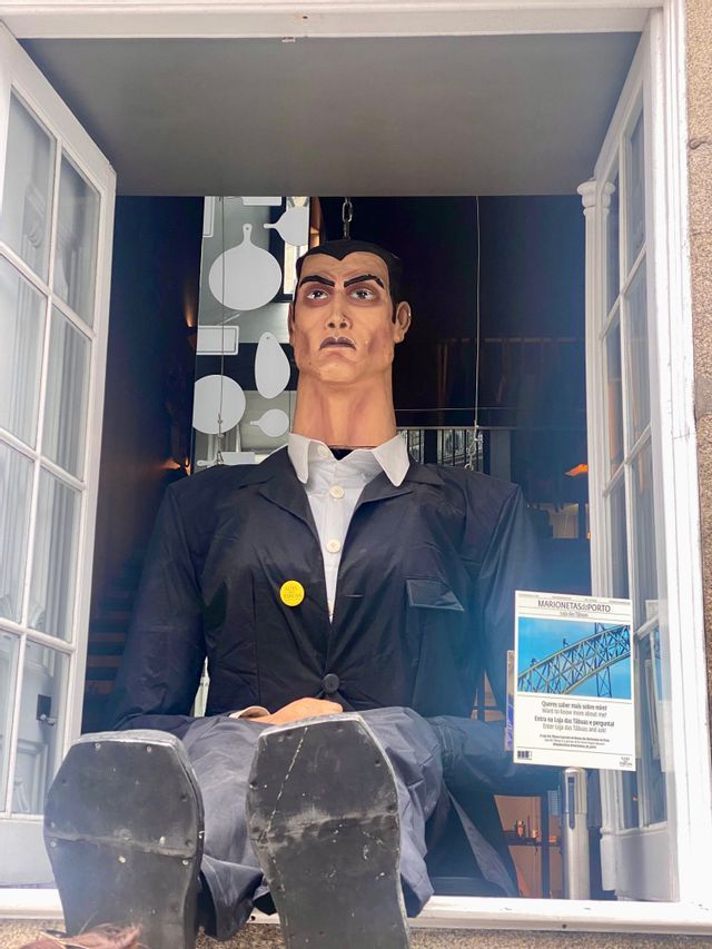 Una marioneta grande, de aspecto humano, con una chaqueta negra, sentada en el alféizar de una ventana abierta y con los pies en alto.