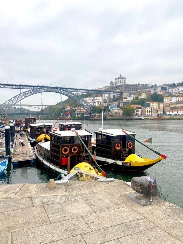 Barcos tradicionales atracados en un muelle de un río, frente a un gran puente metálico y una ciudad en la ladera bajo un cielo nublado.