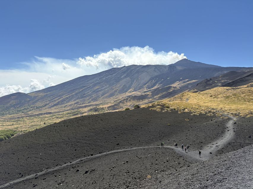 Un viaggio di gruppo WeRoad fa trekking su un sentiero che attraversa un oscuro paesaggio vulcanico, con un vulcano fumante sullo sfondo sotto un cielo azzurro.