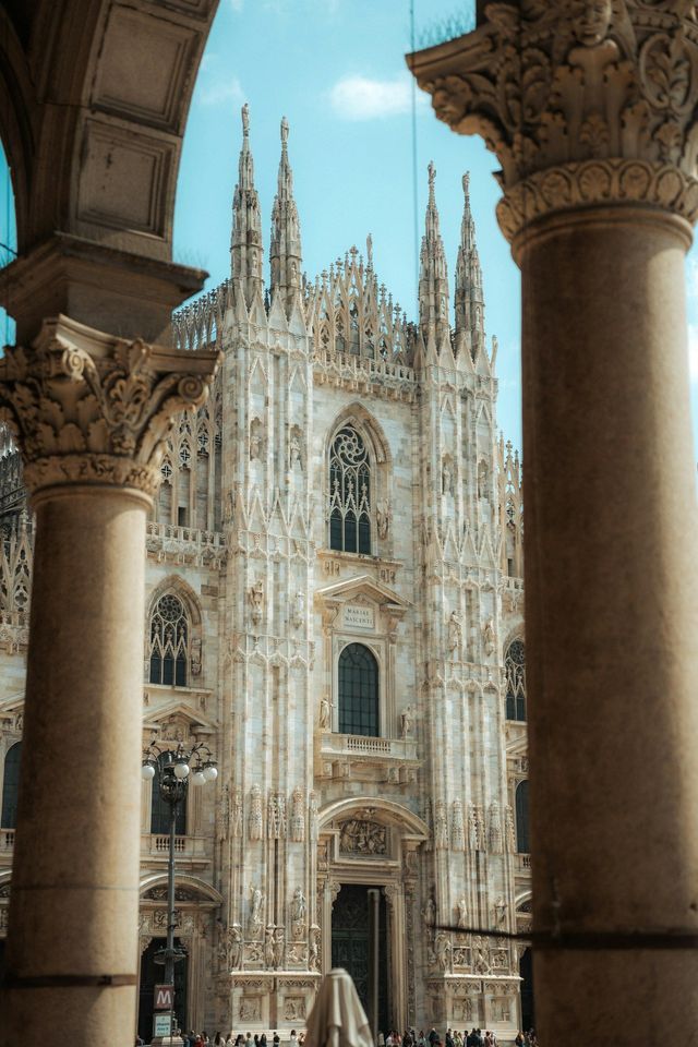 The ornate white facade of a Gothic cathedral with numerous spires, framed between two large, carved stone columns.