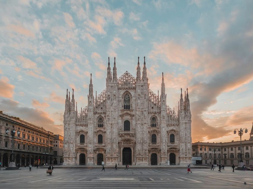 The front facade of an ornate, white Gothic cathedral with numerous spires, viewed from across a large public square at sunset.