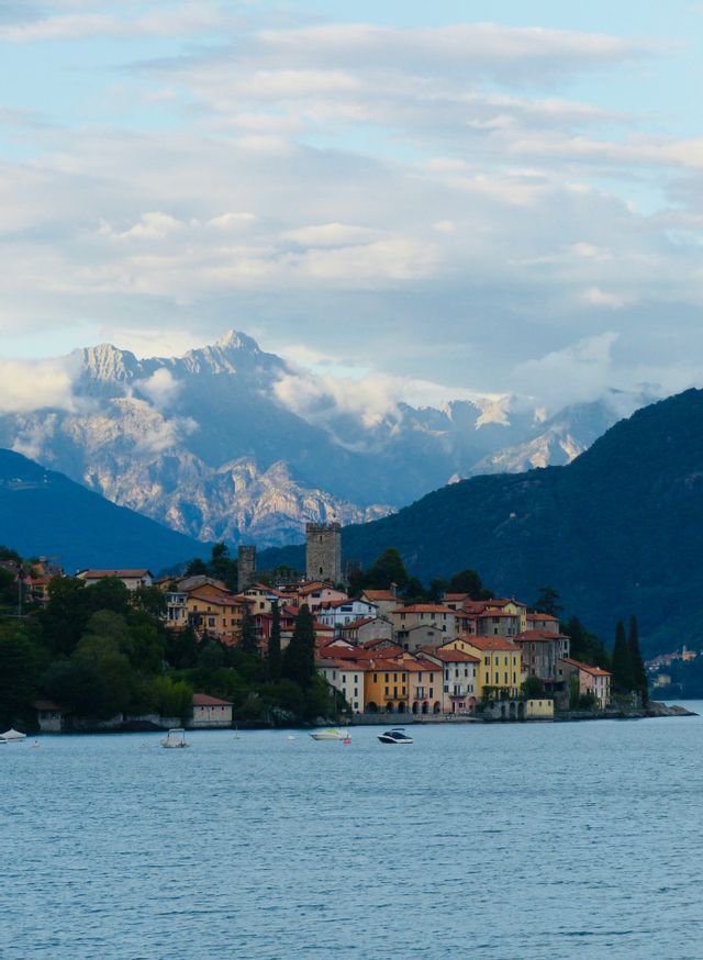 A colorful lakeside village with a stone tower is framed by large mountains under a partly cloudy sky.