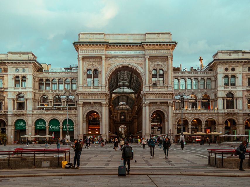 People walk across a large paved plaza in front of a grand, ornate building with a central archway under an overcast sky.