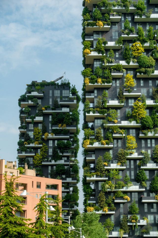 Two modern high-rise buildings functioning as vertical forests, with trees and plants growing on every balcony against a blue sky.