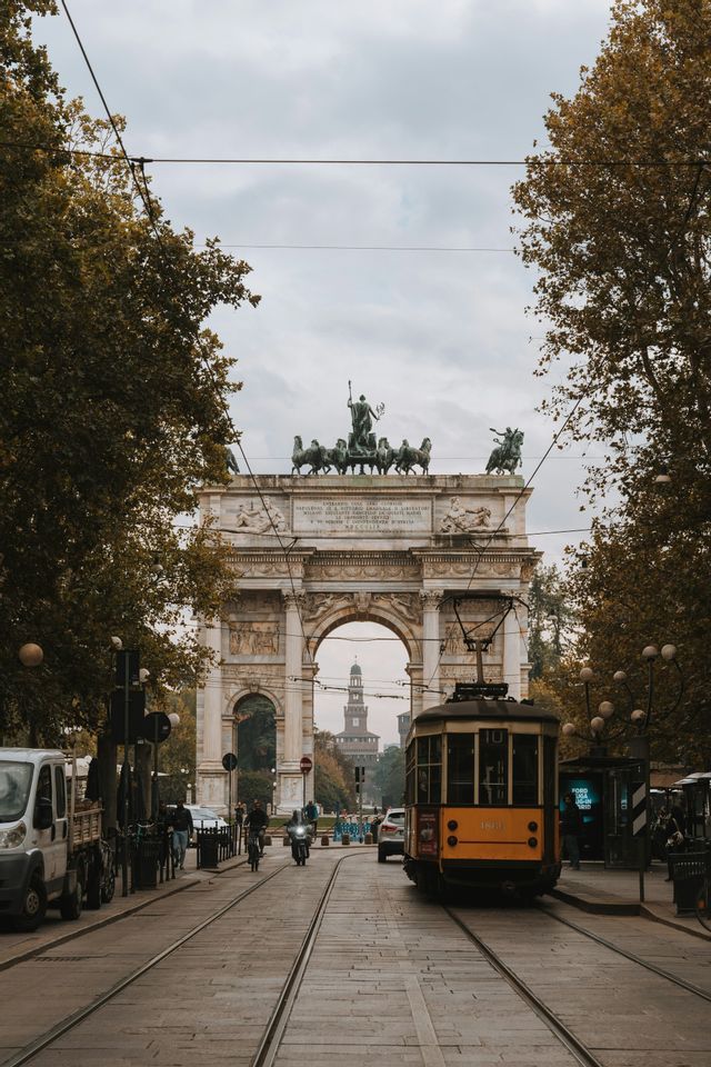 A yellow tram on tracks on a city street leading to a large triumphal arch, with a castle visible in the background.