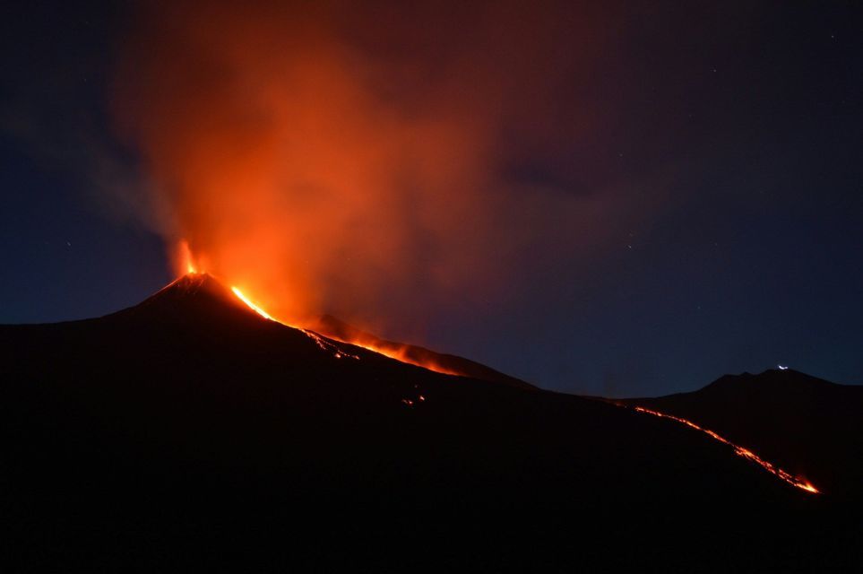 Un vulcano in eruzione di notte, con lava incandescente arancione che scende lungo il pendio e fumo che si innalza nel cielo stellato.