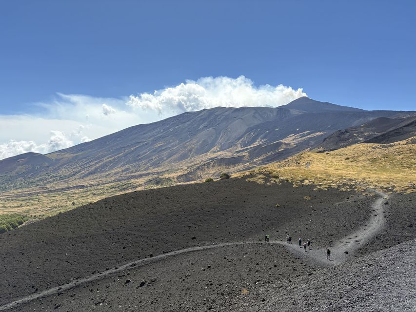 Un viaggio di gruppo WeRoad fa un'escursione su un sentiero tortuoso sui pendii rocciosi e scuri di un grande vulcano fumante.