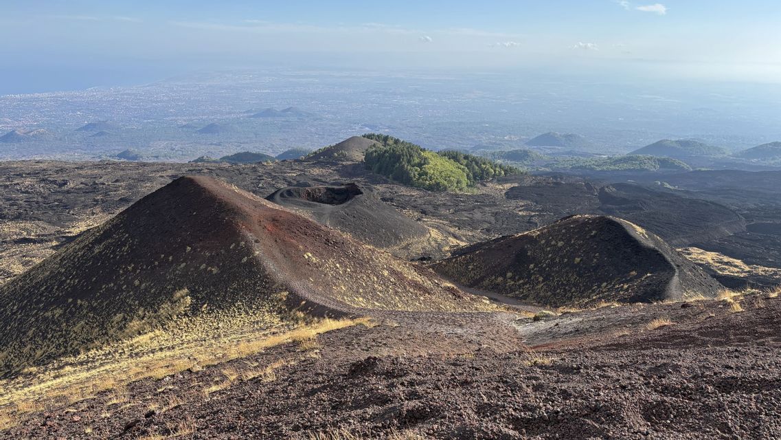 Una vista dall'alto di un vasto paesaggio vulcanico con crateri scuri, che si affaccia su una lontana città costiera sotto un cielo azzurro e limpido.